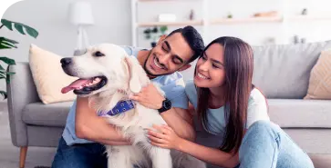 Uma mulher e um homem brancos junto a um cão. Eles vestem roupas claras e estão no chão, próximos de um sofá na sala de estar.