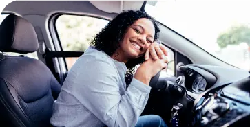 Mulher negra no banco do motorista de seu carro. Ela veste camisa branca e sorri com mãos e rosto apoiados no volante.