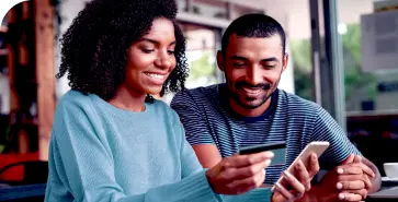 Duas pessoas negras conversam. A mulher veste sueter azul e mostra ao homem seu Cartão BV e a tela de um smartphone.